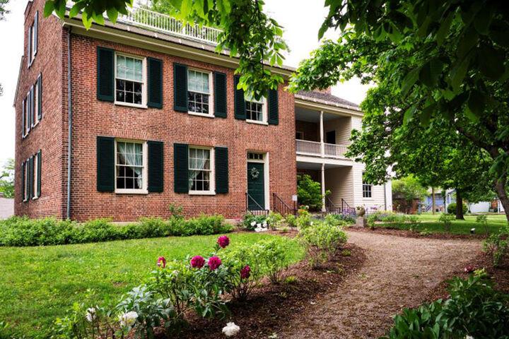 The Wylie House Museum, an old brick, two-story home, is shown surrounded by grass, bushes, flowers, and trees.